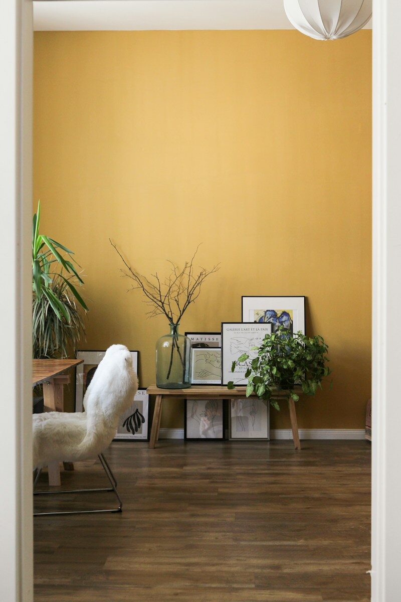 A wooden bench with framed art and plants against a mustard yellow wall